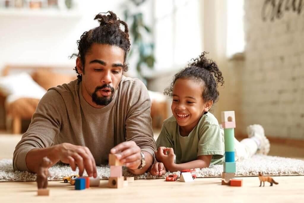 Father and daughter lying on floor of living room playing with toy blocks and toy cars
