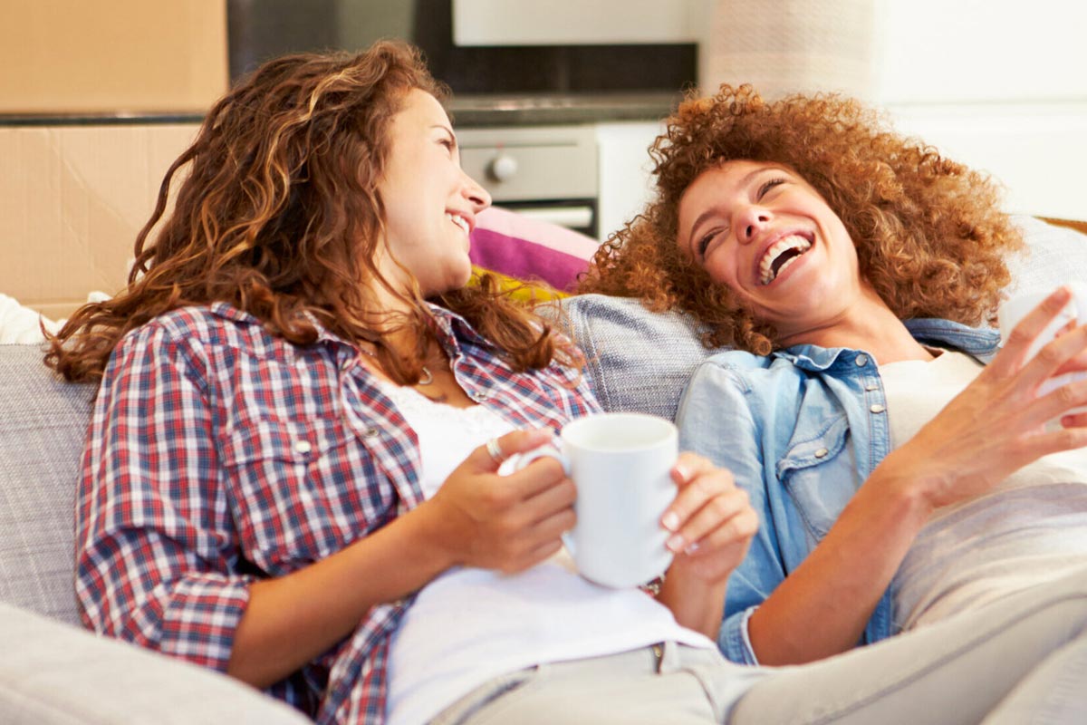 Two women sitting on couch, laughing and holding coffee mugs